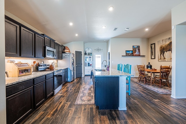 kitchen with dark wood-type flooring, a center island with sink, a sink, appliances with stainless steel finishes, and light stone countertops