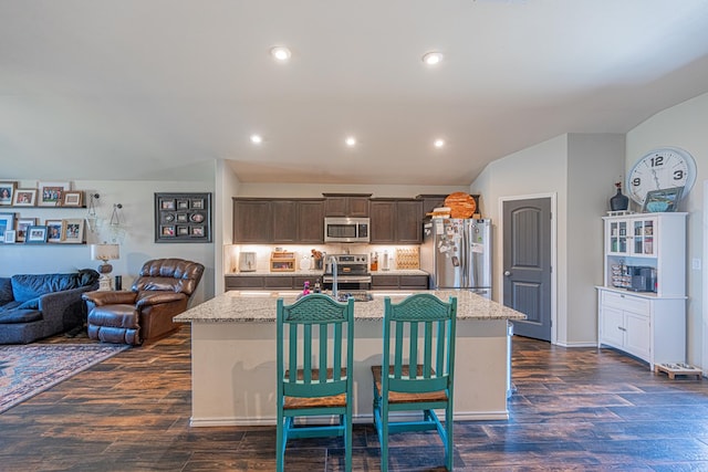 kitchen with dark brown cabinets, light stone countertops, an island with sink, appliances with stainless steel finishes, and dark wood-style floors