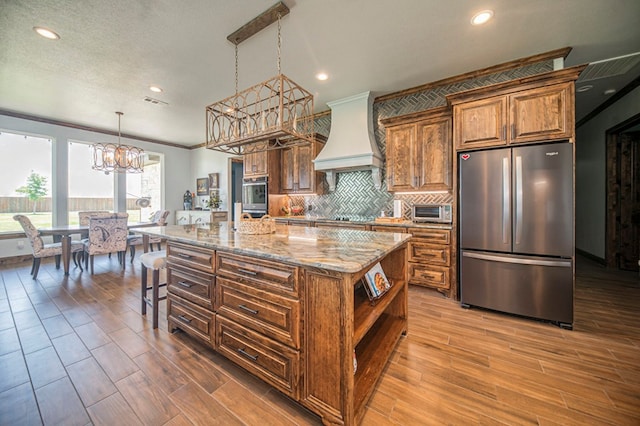 kitchen with pendant lighting, custom exhaust hood, an inviting chandelier, an island with sink, and stainless steel appliances