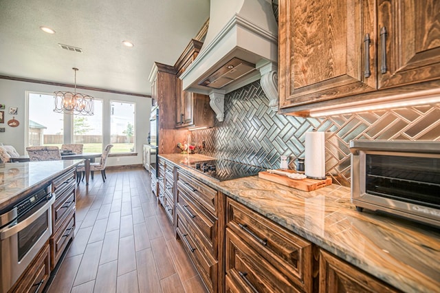 kitchen featuring backsplash, premium range hood, light stone counters, black cooktop, and a chandelier