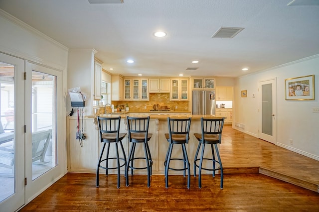 kitchen with stainless steel fridge, a peninsula, glass insert cabinets, wood finished floors, and backsplash