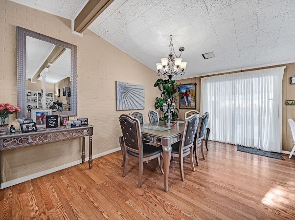 dining space with a notable chandelier, lofted ceiling with beams, wood-type flooring, and brick wall