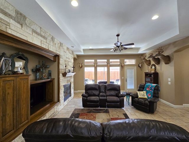 living room featuring a raised ceiling, a ceiling fan, a stone fireplace, and stone tile floors