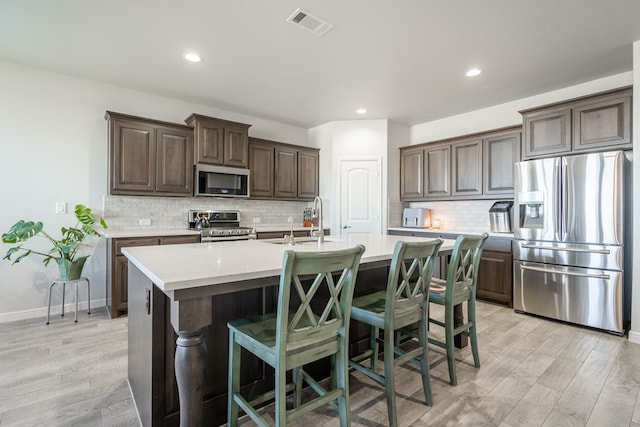 kitchen with dark brown cabinetry, tasteful backsplash, light hardwood / wood-style flooring, an island with sink, and appliances with stainless steel finishes