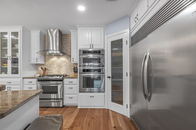 kitchen featuring hardwood / wood-style floors, dark stone counters, wall chimney range hood, appliances with stainless steel finishes, and white cabinetry