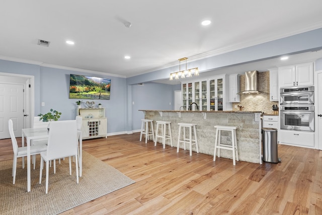 kitchen featuring white cabinetry, wall chimney exhaust hood, double oven, decorative light fixtures, and light wood-type flooring