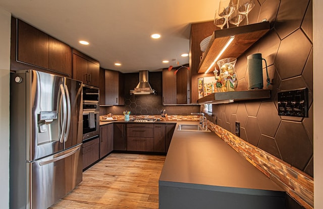 kitchen with dark brown cabinetry, stainless steel appliances, sink, wall chimney range hood, and light hardwood / wood-style flooring