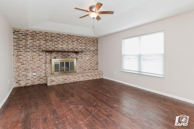 unfurnished living room featuring a raised ceiling, a brick fireplace, dark hardwood / wood-style floors, ceiling fan, and brick wall