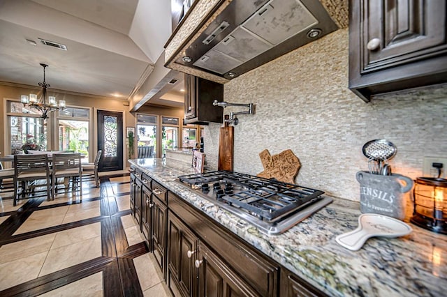 kitchen with dark brown cabinetry, stainless steel gas cooktop, wall chimney exhaust hood, and a notable chandelier