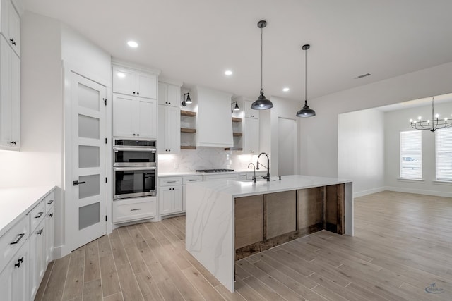 kitchen with white cabinets, light hardwood / wood-style floors, stainless steel double oven, and an island with sink