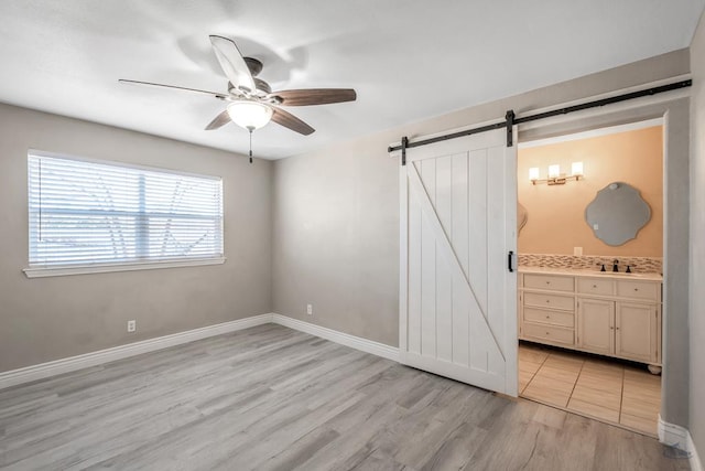 unfurnished bedroom with ensuite bathroom, sink, ceiling fan, a barn door, and light hardwood / wood-style floors