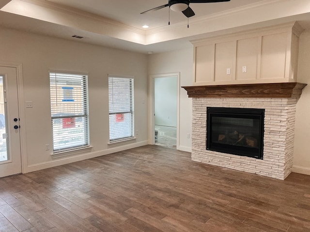 unfurnished living room featuring a raised ceiling, ceiling fan, crown molding, hardwood / wood-style floors, and a stone fireplace