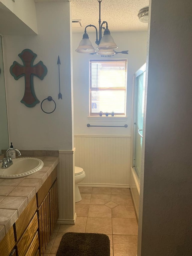 full bath featuring a textured ceiling, tile patterned flooring, toilet, vanity, and wainscoting