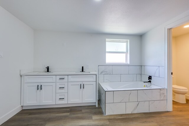 full bath featuring toilet, wood finished floors, a garden tub, double vanity, and baseboards