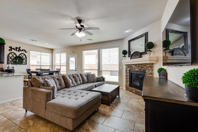 living room with a ceiling fan, visible vents, a stone fireplace, and a textured ceiling