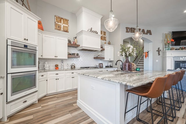 kitchen with pendant lighting, an island with sink, white cabinets, white double oven, and backsplash