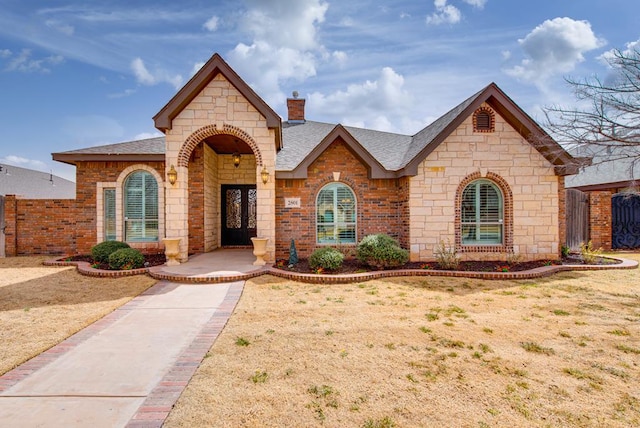 french provincial home with a shingled roof, a chimney, fence, french doors, and brick siding