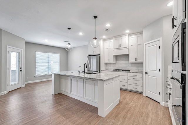 kitchen with pendant lighting, appliances with stainless steel finishes, white cabinetry, a kitchen island with sink, and light stone countertops