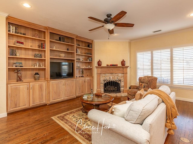 living room with visible vents, dark wood-style flooring, ceiling fan, a stone fireplace, and crown molding