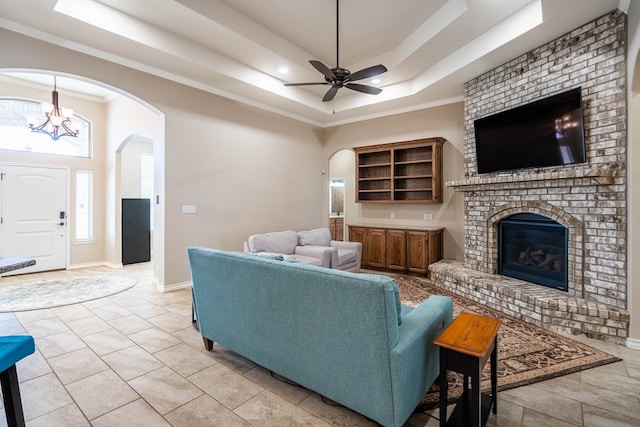 living area with ceiling fan with notable chandelier, a fireplace, baseboards, ornamental molding, and a raised ceiling