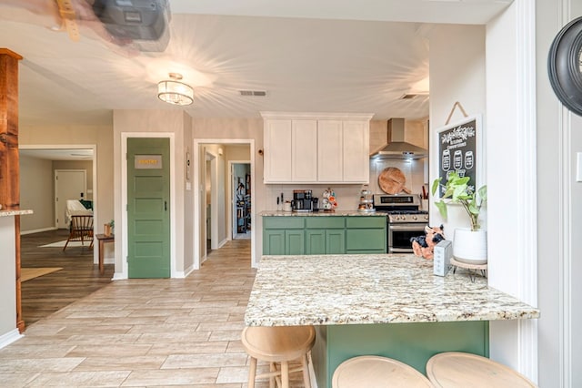 kitchen featuring a breakfast bar, white cabinets, green cabinets, wall chimney range hood, and stainless steel stove