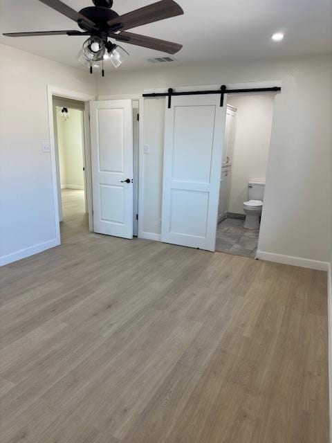 unfurnished bedroom with ceiling fan, a barn door, light wood-type flooring, and connected bathroom