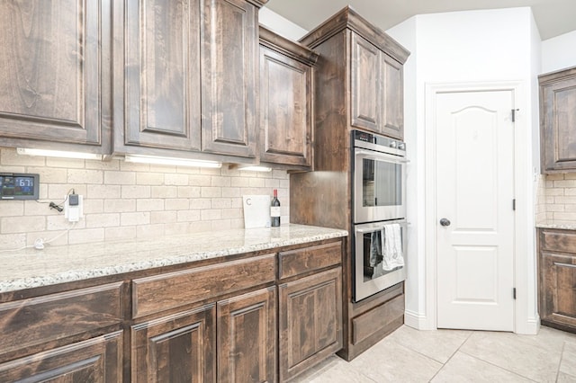 kitchen featuring double oven, backsplash, dark brown cabinets, and light stone countertops