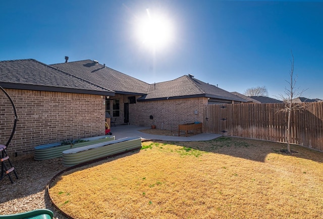 rear view of house with roof with shingles, brick siding, a patio, a lawn, and fence