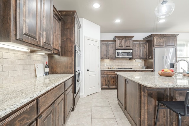 kitchen featuring stainless steel appliances, tasteful backsplash, light tile patterned flooring, dark brown cabinetry, and light stone countertops