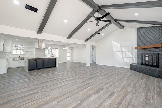 unfurnished living room featuring light wood-style flooring, a brick fireplace, a sink, ceiling fan, and beamed ceiling