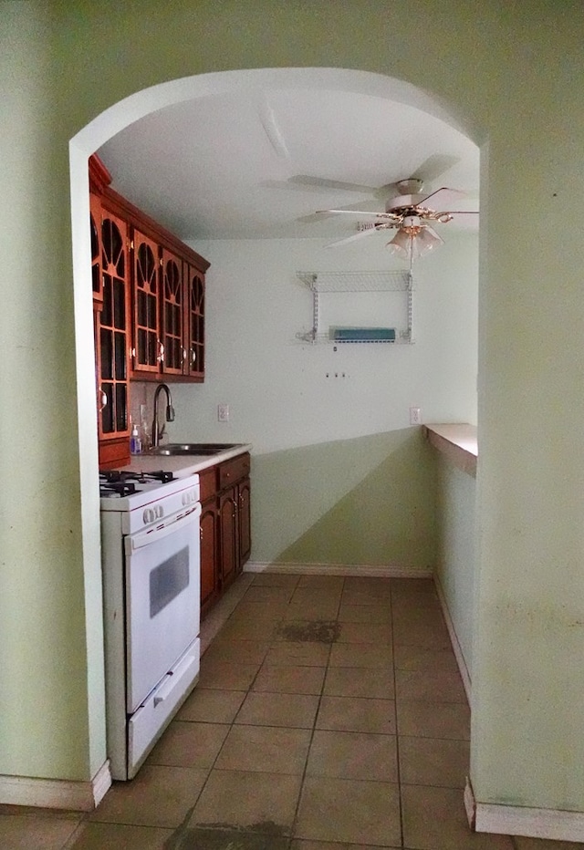 kitchen with white gas range, a ceiling fan, light tile patterned floors, a sink, and glass insert cabinets