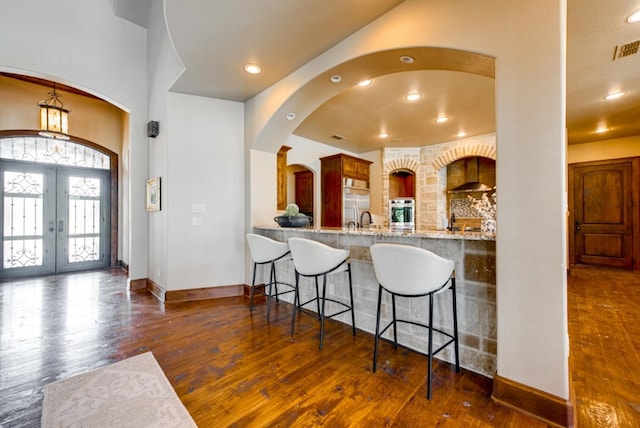 kitchen featuring oven, french doors, light stone countertops, wall chimney range hood, and dark wood-type flooring