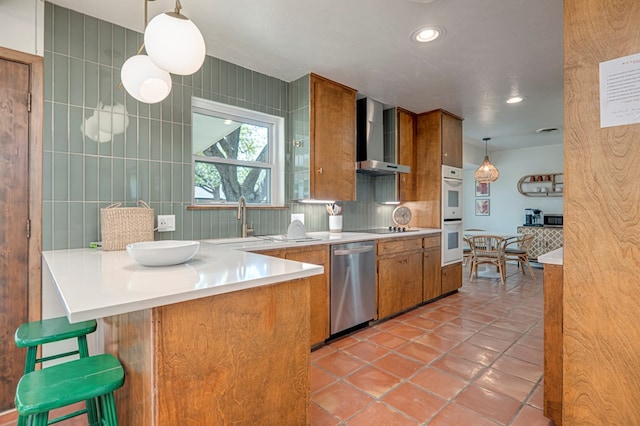 kitchen with wall chimney range hood, decorative backsplash, stainless steel dishwasher, brown cabinets, and a sink