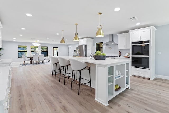 kitchen with pendant lighting, wall chimney range hood, black double oven, light hardwood / wood-style floors, and white cabinetry