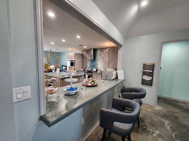 kitchen featuring kitchen peninsula, stainless steel appliances, vaulted ceiling, wall chimney range hood, and a breakfast bar area