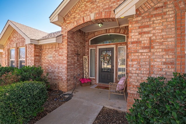 doorway to property featuring covered porch, roof with shingles, and brick siding