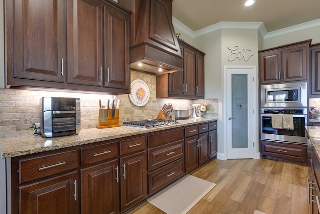 kitchen featuring stainless steel appliances, custom range hood, dark brown cabinets, ornamental molding, and light wood-style floors