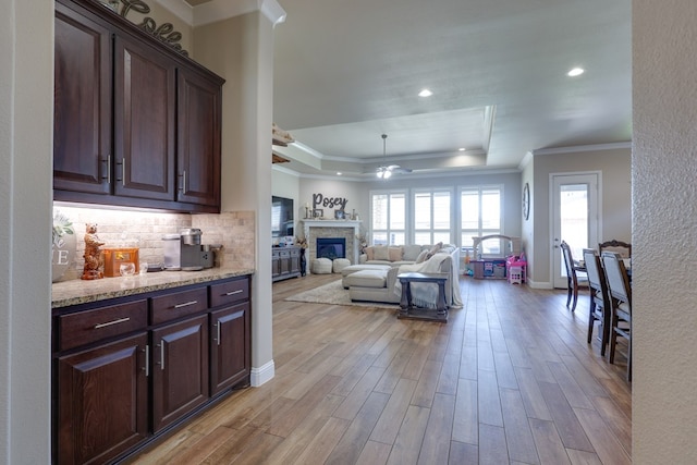 kitchen featuring a raised ceiling, ceiling fan, ornamental molding, a glass covered fireplace, and light wood-type flooring
