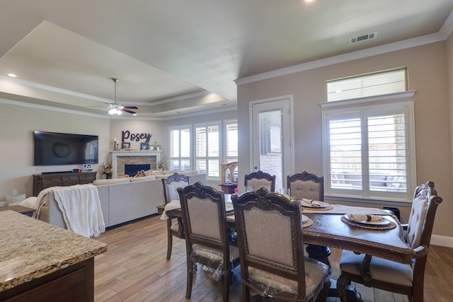 dining room featuring a tray ceiling, ceiling fan, light wood-style floors, ornamental molding, and a fireplace