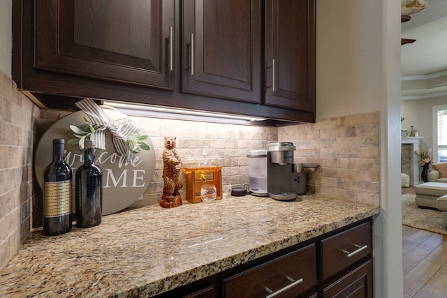 kitchen with dark brown cabinetry, wood finished floors, tasteful backsplash, light stone countertops, and ornamental molding