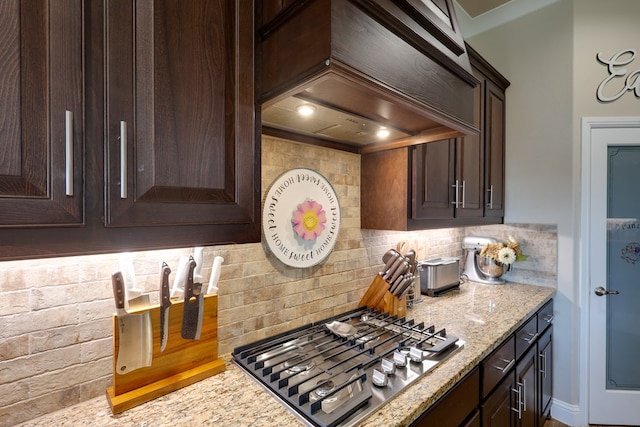 kitchen with stainless steel gas stovetop, dark brown cabinets, backsplash, light stone counters, and premium range hood