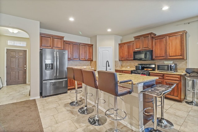 kitchen with light stone counters, backsplash, a breakfast bar area, a kitchen island with sink, and black appliances