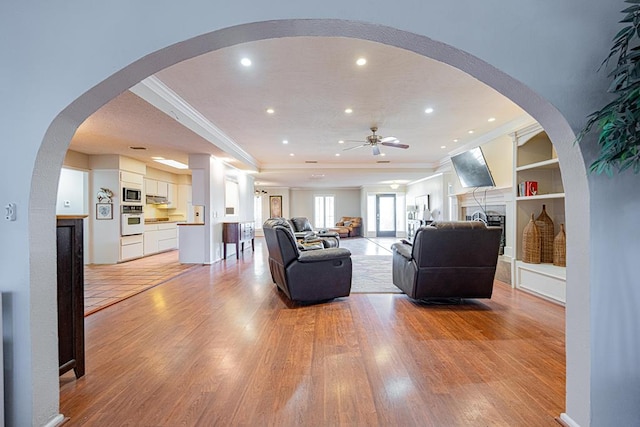 living room featuring built in shelves, ornamental molding, a raised ceiling, and light wood-type flooring