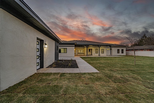 back of house at dusk with a patio area, stucco siding, fence, and a lawn
