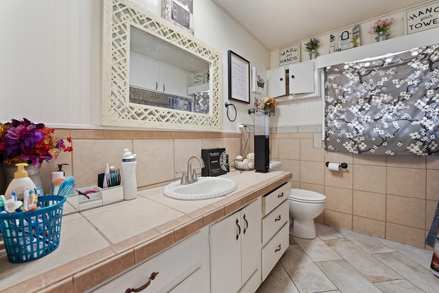 full bath with tile patterned flooring, tile walls, toilet, wainscoting, and vanity