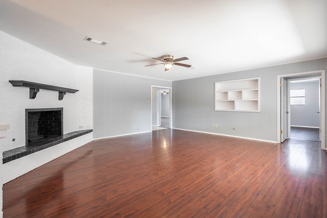 unfurnished living room with built in features, dark wood finished floors, visible vents, a ceiling fan, and a brick fireplace