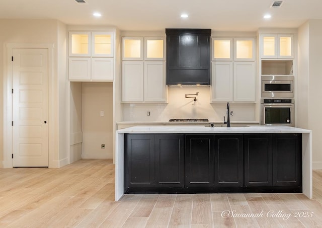 kitchen featuring wall chimney range hood, sink, appliances with stainless steel finishes, white cabinetry, and an island with sink