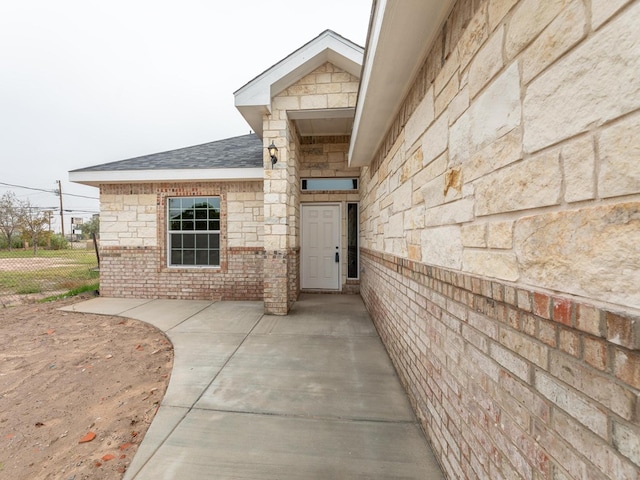 entrance to property featuring brick siding, fence, and roof with shingles