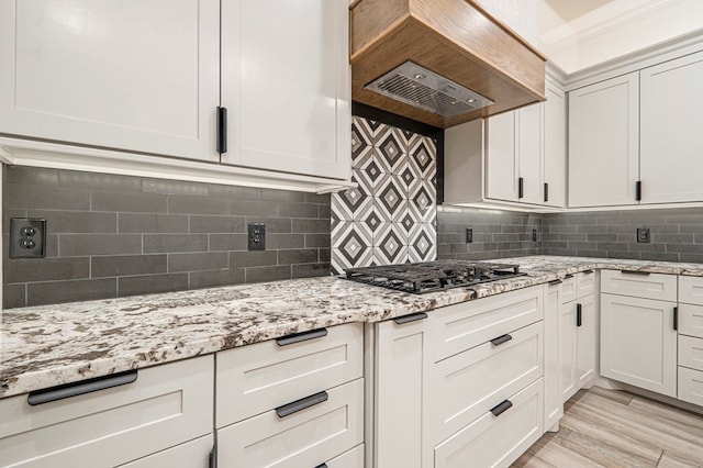 kitchen with light stone counters, custom range hood, white cabinets, stainless steel gas stovetop, and backsplash