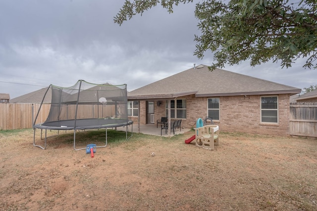 rear view of house featuring a trampoline, a shingled roof, brick siding, a fenced backyard, and a patio